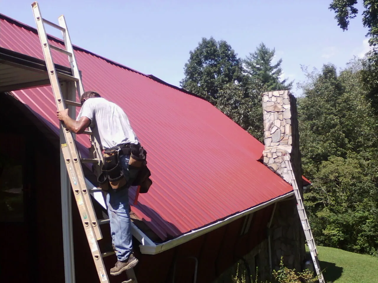 Roofer on a ladder beside a red metal roof and stone chimney — Soffit & Fascia Repair by Roofing Brothers in Rodeo
